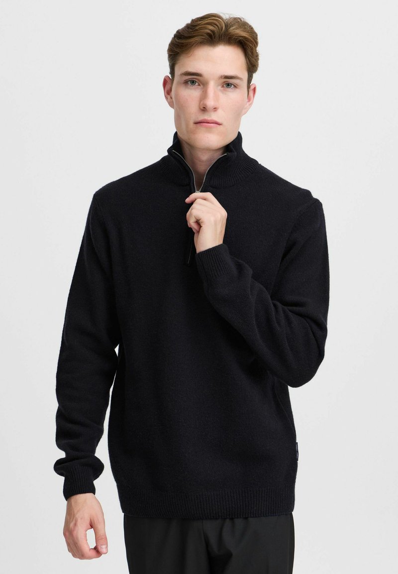 Pull à col zippé noir en tissu doux, avec des poignets et un ourlet côtelés, coupe décontractée, col standard et étiquette discrète sur le côté.