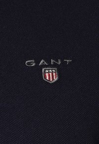 Marineblaues Gewebe mit strukturiertem Gewebe. Verfügt über gesticktes "GANT"-Logo und Wappen mit roten und weißen Streifen. Subtile, klassische Designelemente.