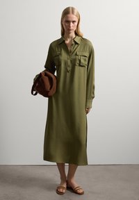 Olivgrünes, langärmeliges Hemdkleid mit zwei Brusttaschen, seitlichen Schlitzen und glatter Textur. Das Model hält eine braune Tasche und trägt Slide-Sandalen.