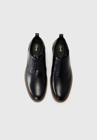 Zapatos de cuero negro con un acabado suave, punta redonda, diseño con cordones y adornos de suela en un marrón claro que contrastan. En el interior presentan la marca Clarks.