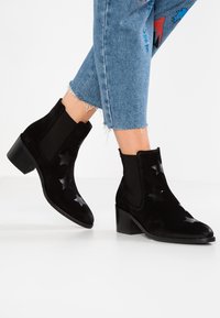 Stivaletti neri in suede con fori a forma di stella, pannelli laterali elasticizzati e tacco a blocco. Presentano una superficie liscia e un design con punta appuntita.