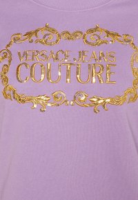 Jasnofioletowa bluza z nadrukiem w stylu barokowym wykonanym ze złotych cekinów, z widocznym napisem „VERSACE JEANS COUTURE” w centralnej części.