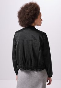 Veste bomber noire légère avec taille et poignets élastiques, présentant une texture lisse et légèrement brillante ainsi qu'un design simple.