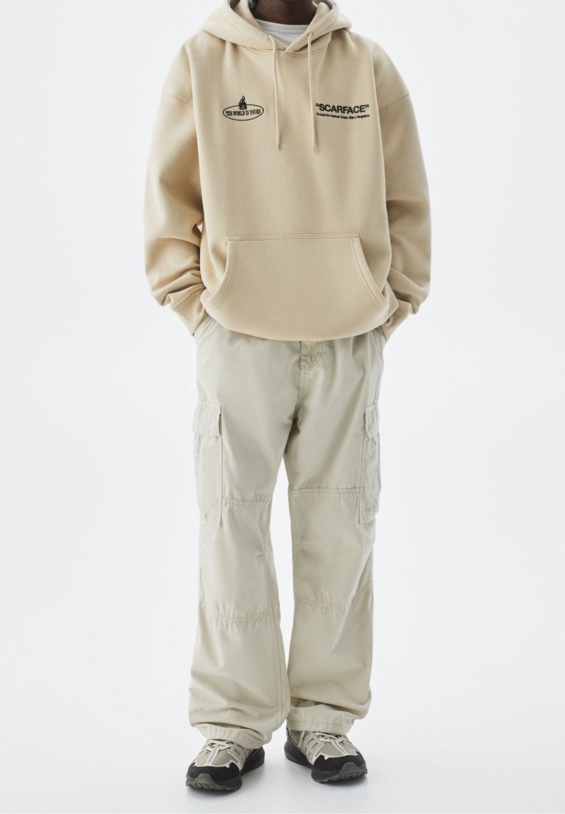 Personne porte un sweat à capuche beige avec le texte « SCARFACE », un pantalon cargo clair et des baskets noir et beige, debout les mains dans les poches.