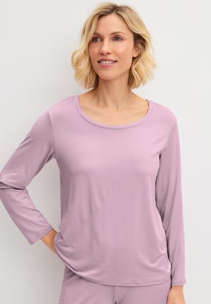 Femme aux cheveux blonds courts portant une chemise à manches longues violet clair et un pantalon assorti, posant avec une main sur la hanche.