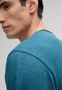 Türkises Rundhals-T-Shirt aus strukturiertem Stoff. Nahaufnahme der Schulter und des Halsausschnitts, die einen lässigen Schnitt und glatte Nähte zeigen.