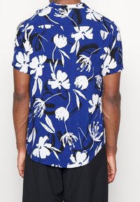 Lyhyet hihnat takki sininen kukkakuviolla valkoisena ja mustana. Varustettu leirikauluksella ja kaarevalla helmalla takana. Kevyt kangas.