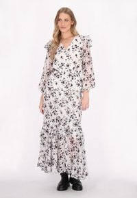 Letende maxikjole i let, gennemsigtig stof med hvid bund og sort blomsterprint. Har voluminøse ærmer og lagdelt skirt. Sorte støvler iført.