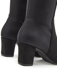 Botines negros de tacón alto que presentan un material superior liso con un tacón de ante texturizado. El diseño incluye una puntera redondeada y un tacón robusto.