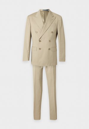 Homme portant un costume beige croisé avec une chemise blanche et des baskets blanches, debout devant un fond blanc uni.