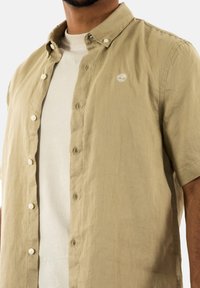 Camicia a maniche corte di color beige chiaro con bottoni, realizzata in tessuto testurizzato, con un logo sul petto e chiusure a bottone standard.