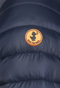 Donkerblauwe pufferjack met een geborduurde textuur en een oranje cirkelvormige patch met een eend en de tekst "SAVE THE DUCK".