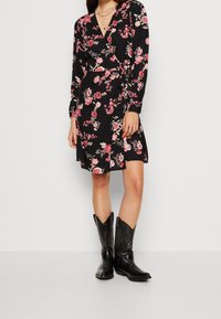 Robe noire à manches longues, motif floral en rose et blanc, décolleté en V, taille cintrée, associée à des bottes de cowboy en cuir noir.