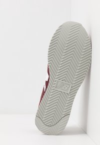 Semelle de basket rouge avec une partie inférieure en caoutchouc gris texturé. Présente un logo "N" bien visible et un motif de bande de roulement en chevrons pour une meilleure adhérence.