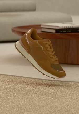 Barna sportcipő lebeg egy bézs szőnyeg felett egy modern nappaliban, háttérben kerek fa dohányzóasztal és könyvek.