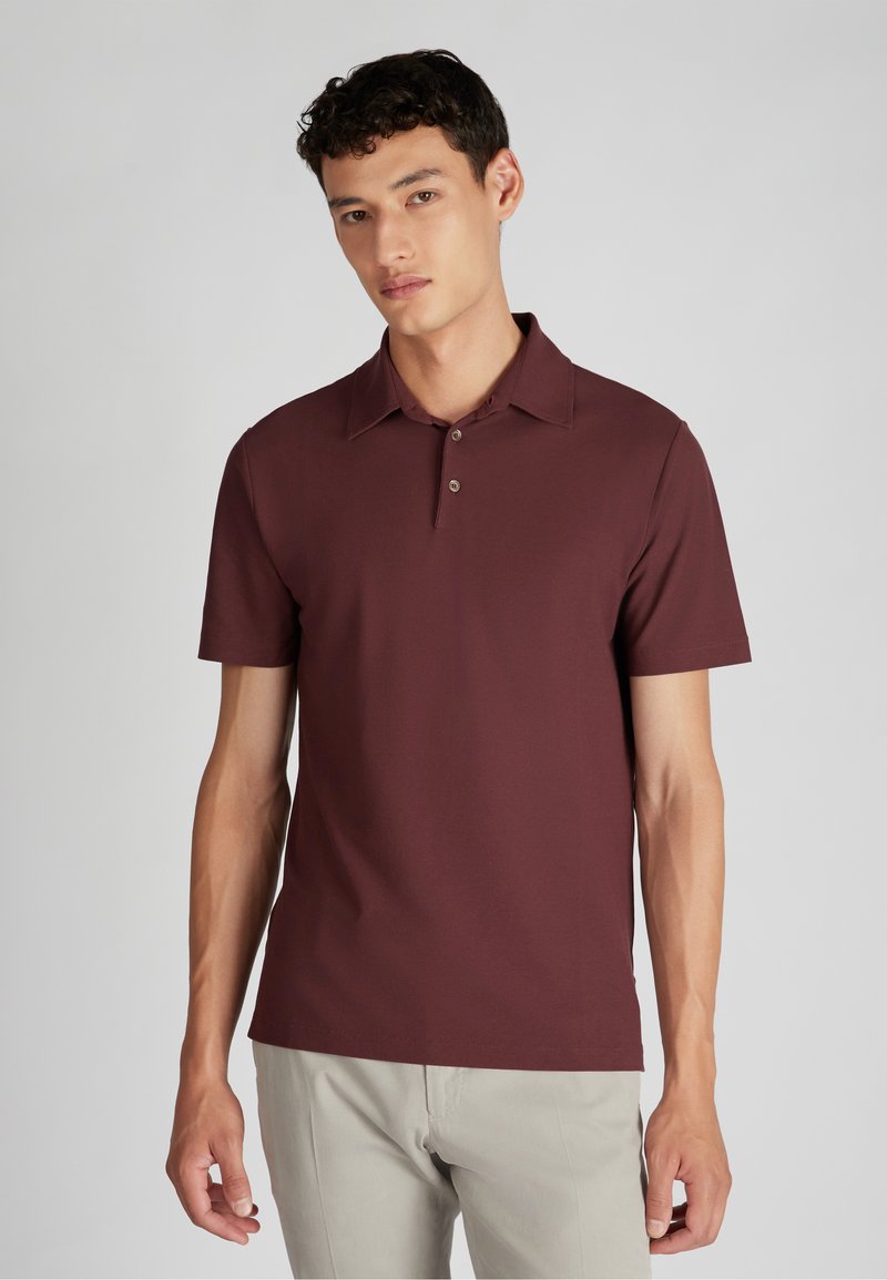 Jongeman in een lichtgrijze achtergrond, gekleed in een kortärmige bordeauxrode polo en lichtbeige broeken.