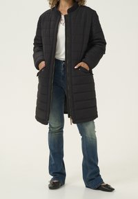 Abrigo largo negro acolchado con cuello acanalado, costuras horizontales, cierre de cremallera y bolsillos laterales, combinado con vaqueros de campana azules y zapatos planos negros.