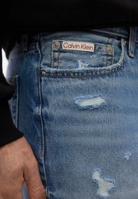 Κοντινό πλάνο σε μπλε τζιν παντελόνι με ετικέτα Calvin Klein στην μπροστινή τσέπη και ένα χέρι που ακουμπάει δίπλα στην τσέπη.