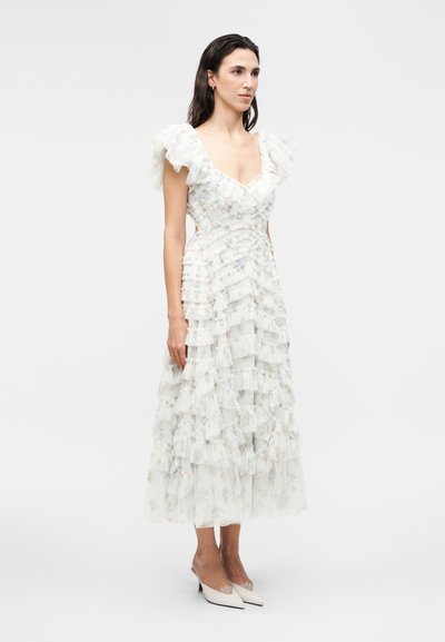 Γυναίκα που φοράει λευκό φόρεμα μίντι με λουλουδάτο σχέδιο, στρωματωτές βολάν και φουσκωτά μανίκια, με λευκές μυτερές γόβες σε μονόχρωμο φόντο.