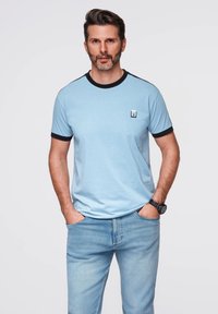 Světlá modrá bavlněná tričko s černým žebrovaným límcem a lemy rukávů, s malou logovou nášivkou. Doplněno vybledlými modrými džínami.