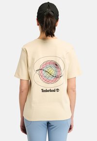 Camiseta de manga corta beige con un diseño de globo multicolor y texto en la parte posterior, con un logo negro en la parte inferior.