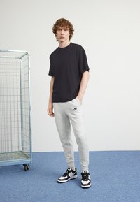 Un giovane in piedi con una mano in tasca, indossando una maglietta nera, pantaloni sportivi grigi della Nike e sneakers bianco e nere, accanto a un carrello di metallo per lo stoccaggio.