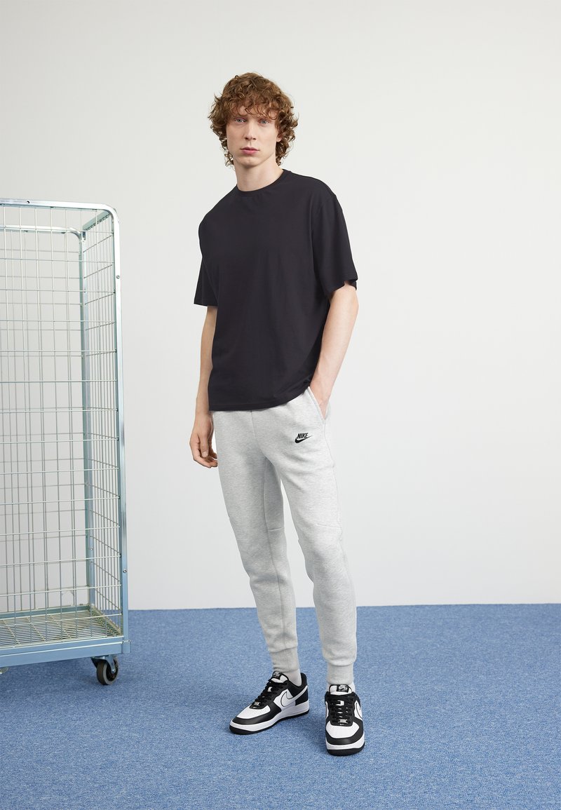 Junger Mann steht mit einer Hand in der Tasche, trägt ein schwarzes T-Shirt, graue Nike-Sweatpants und schwarz-weiße Sneaker, neben einem Metalllagerwagen.