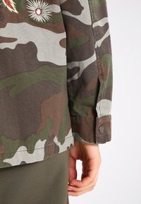 Skjorta med kamouflagemönster i gröna och bruna toner, med broderade detaljer och knäppt ärmslut. Materialet ser texturerat och vävt ut.