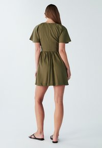 Olive zelena kratka obleka s kratkimi rokavi, prilegajočim zgornjim delom in razširjenim krilom. Ima zadrgo na hrbtni strani in gladko, lahko tkanino.