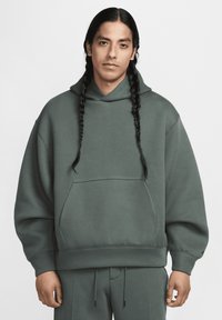 Homem com cabelo trançado longo, vestindo um moletom verde oversized e calças a condizer, de pé contra um fundo claro e simples.