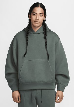 Mann mit langen geflochtenen Haaren, der einen übergroßen grünen Hoodie und passende Hosen trägt, steht vor einem schlichten hellen Hintergrund.