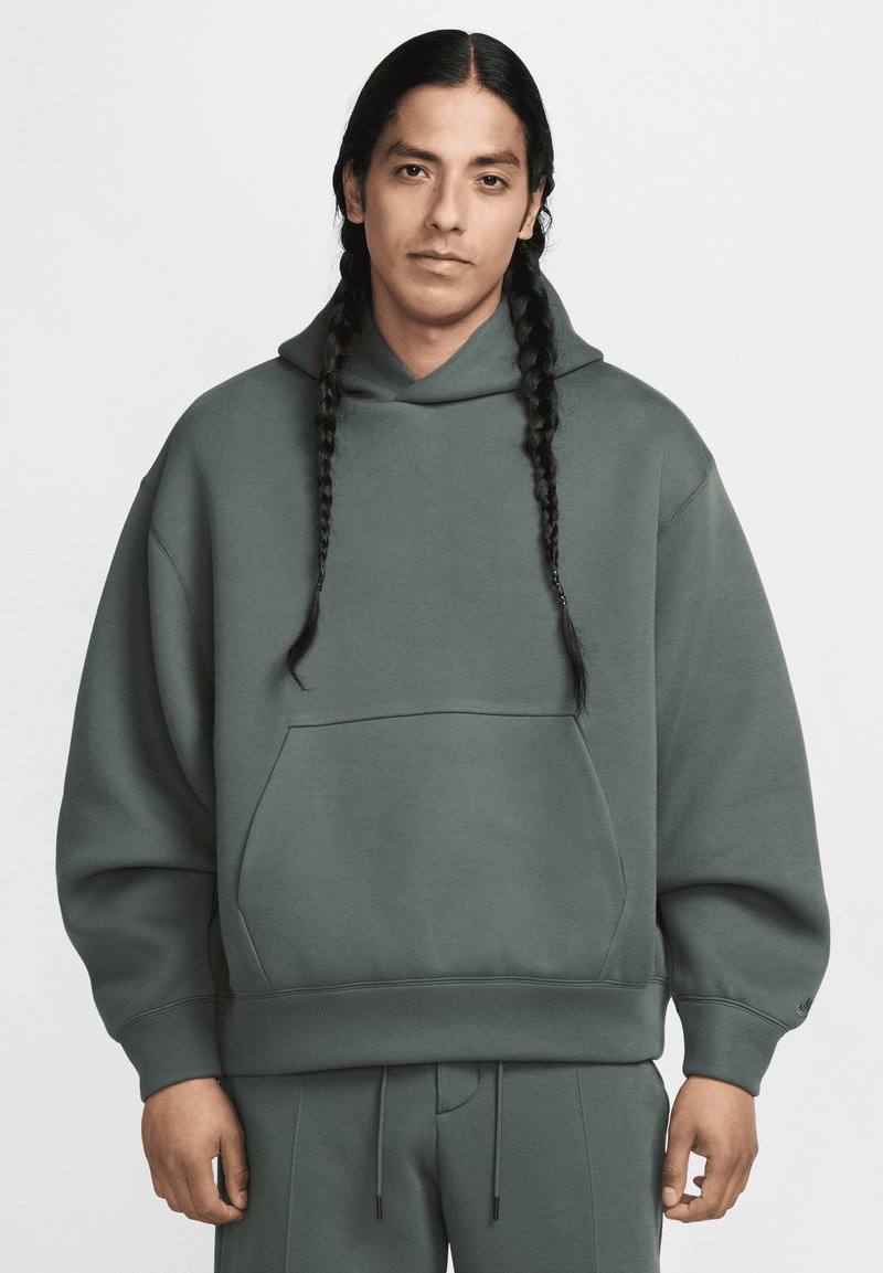 Homem com cabelo trançado longo, vestindo um moletom verde oversized e calças a condizer, de pé contra um fundo claro e simples.