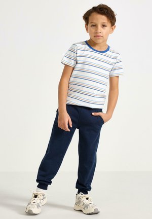 Pojkars outfit med en vit t-shirt med blå ränder och navyblå joggerbyxor, kombinerat med beige och grå sneakers.