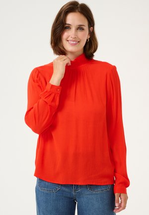 Red long-sleeve blouse with a high ruffled neckline, pleated front detail, and buttoned cuffs, paired with blue jeans.
