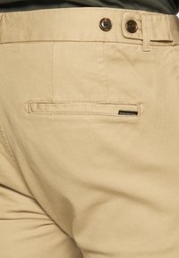 Beige cotton trousers with textured fabric, featuring belt loops and two brown buttons at the waist. Includes a back pocket with logo.