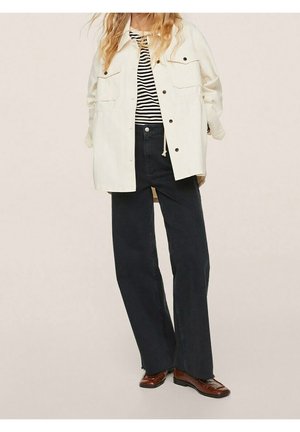 Veste beige clair à boutons avec deux poches poitrine portée sur une chemise à rayures noir et blanc. Associée à un jeans large sombre et des chaussures brunes.