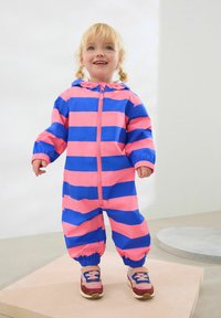 Felroze en blauwe gestreepte regenpak met elastische manchetten en een voorrits. Draagt kleurrijke sneakers en staat op een lichte plaat.