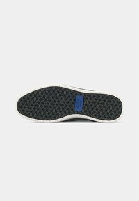 Sola di scarpa in gomma nera con battistrada a motivo diamante, dettaglio della suola intermedia bianca e accento con logo blu vicino all'arco.