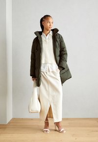 Gevoerde olijfgroene jas, witte gebreide trui, gelaagd wit overhemd, crèmekleurige maxi-jurk met een split, en witte sandaaltjes, terwijl ze een beige tas vasthoudt.
