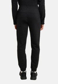 Jogging noir en tissu technique avec une taille élastique, des chevilles resserrées et un tissu doux et texturé, associés à des chaussures de sport noires.