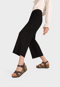 Sandali wedge marroni con cinturini incrociati e fibbia regolabile. Suola texturizzata e soletta in sughero. Sono visibili anche pantaloni cropped neri e una maglietta bianca.