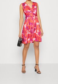 Robe fleurie avec un motif rose et rouge, corsage ajusté et jupe évasée. Portée avec des talons noirs à lanières et un petit sac à main noir.