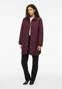 Bordeaux gekleed gewatteerd jasje met een kraag, drukkers en lange mouwen. Gecombineerd met zwarte broek en puntige zwarte schoenen. Fluweelzachte textuur.