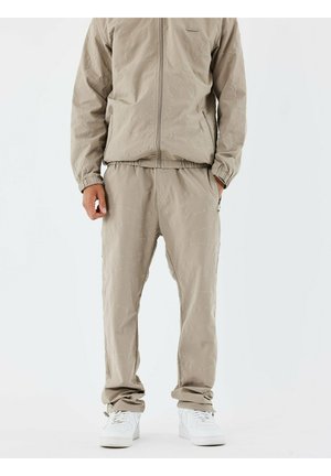 Chándal beige claro con chaqueta con cremallera y pantalones de cintura elástica que presentan patrones de costura sutiles. Combinado con zapatillas blancas.