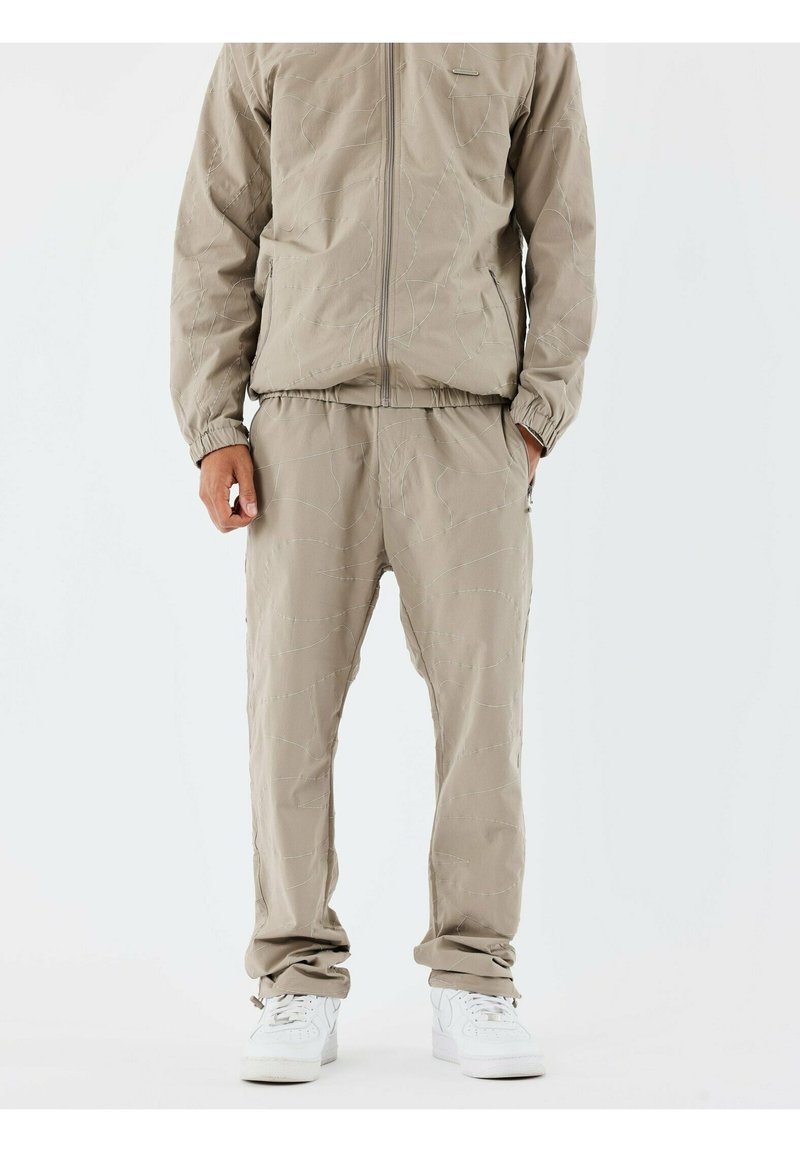 Ensemble de survêtement beige clair avec une veste zippée et un pantalon à taille élastique présentant des motifs de couture subtils. Associé à des baskets blanches.