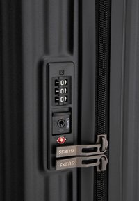 Dettaglio di una valigia rigida nera con lucchetto a combinazione, porta di accesso TSA e doppie cerniere con marchio "GUESS". Superficie texture.