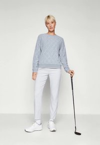 Svetlo siva pulover z diamantnim vzorcem, kombiniran z belimi ožanimi hlačami. Model drži golf palico, obut v bele športne čevlje.