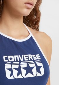 Granatowy bezrękawnik halter z białymi detalami; charakteryzuje się dużym napisem "CONVERSE" i grafiką gwiazdy. Gładka tkanina, dopasowany krój.