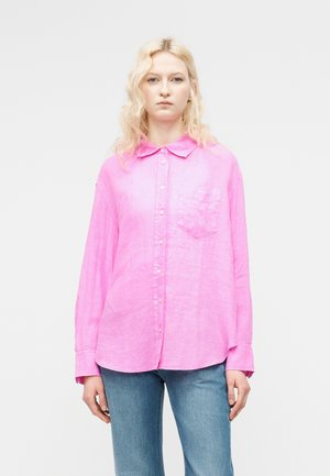 Ξανθιά γυναίκα με κυματιστά μαλλιά, φορώντας ένα χαλαρό, φωτεινό ροζ πουκάμισο με κουμπιά και μπλε τζιν, στέκεται μπροστά σε απλό λευκό φόντο.