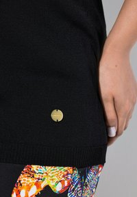 Schwarzes Strickoberteil mit geripptem Saum, featuring ein goldfarbenes Logoschild. Kombiniert mit buntem, gemustertem Stoff, der lebendige, abstrakte Designs präsentiert.
